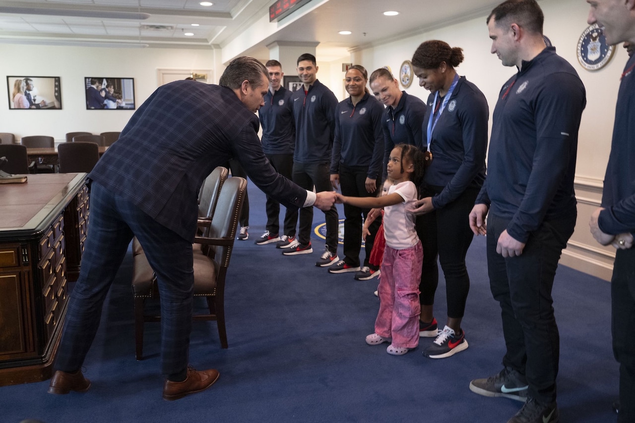 Nearly a dozen people in athletic attire stand in a conference room as a man in business attire shakes hands with a small child.
