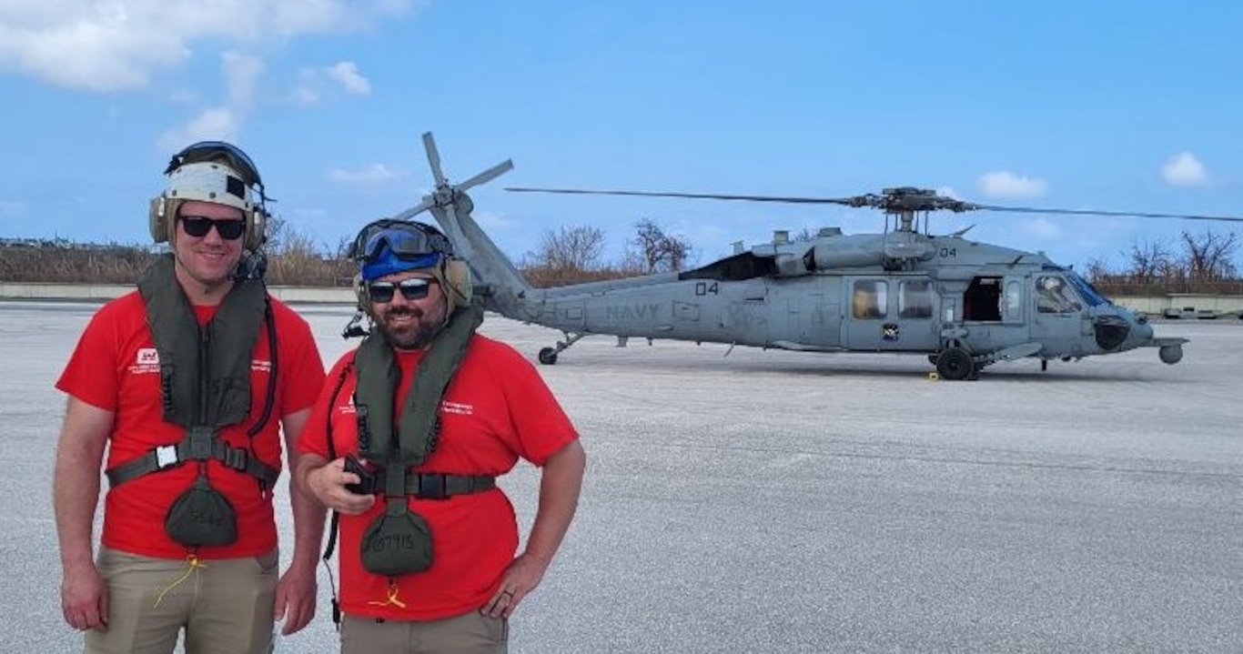 Two men in red shirts stand in front of a Navy helicopter.