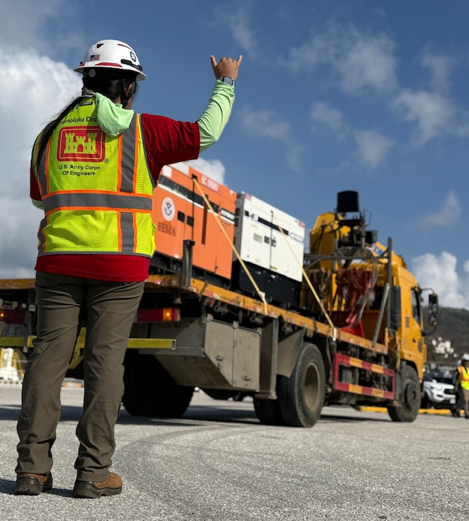 A person wearing a hard hat and a reflective vest, stands on near the back corner of a flat bed truck loaded with generators. The person is waiving their hand guiding the truck as it is backing up.