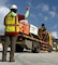 A person wearing a hard hat and a reflective vest, stands on near the back corner of a flat bed truck loaded with generators. The person is waiving their hand guiding the truck as it is backing up.