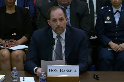 A man in business attire sits at a desk and speaks into a microphone while several people in military dress uniforms and business attire are seated behind him.
