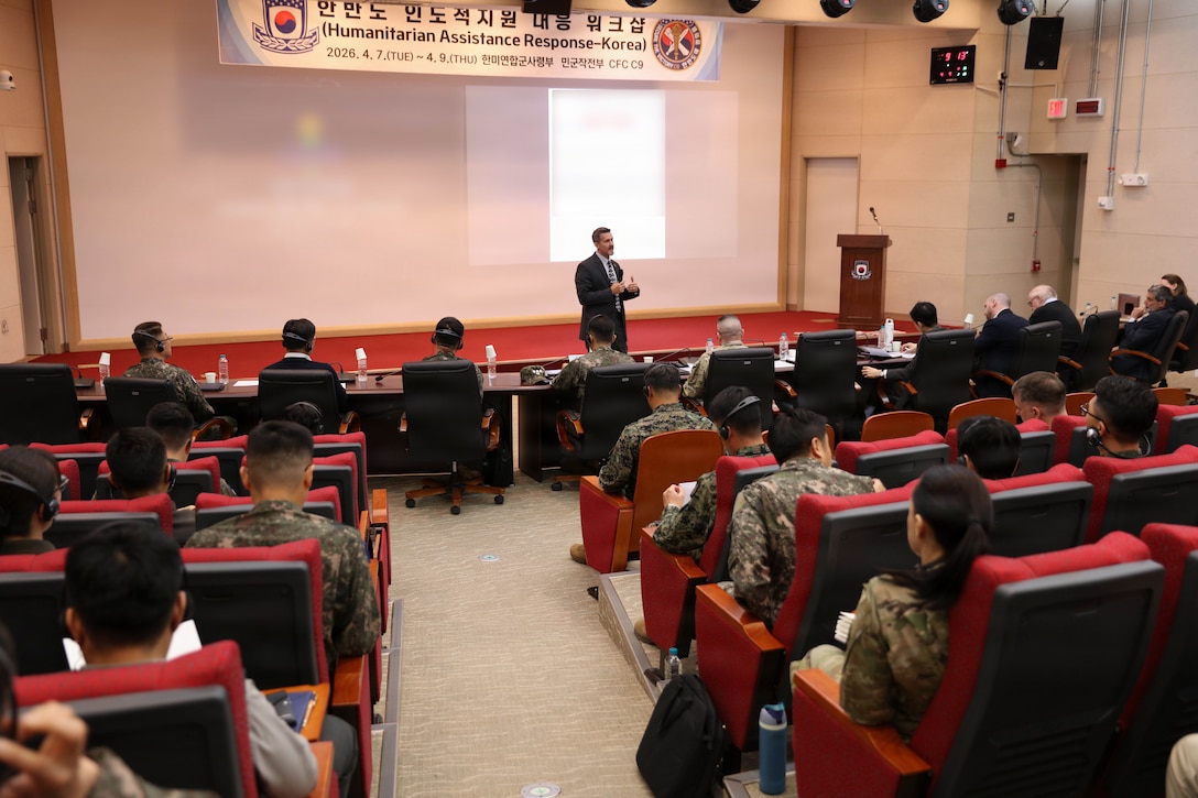 A man in a suit stands on a stage and gives a presentation to an audience seated in a large auditorium. The audience, composed of military personnel in uniform and civilians, is seen from behind, listening to the speaker. A banner above the stage reads "Humanitarian Assistance Response-Korea"