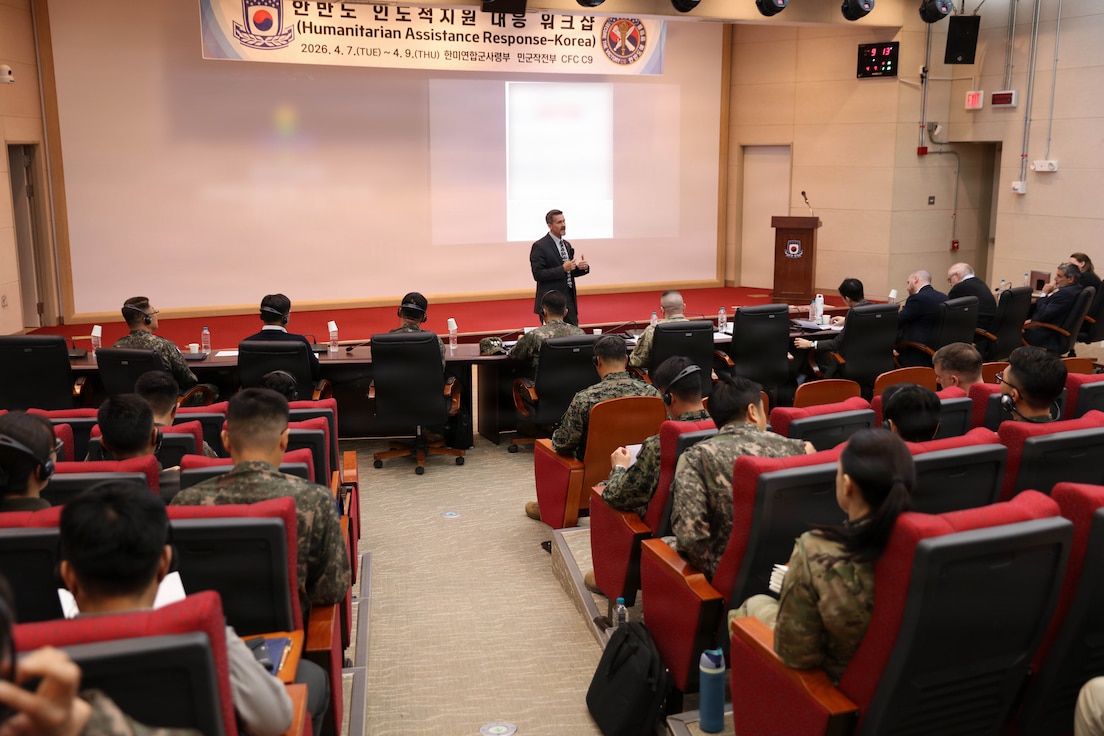 A man in a suit stands on a stage and gives a presentation to an audience seated in a large auditorium. The audience, composed of military personnel in uniform and civilians, is seen from behind, listening to the speaker. A banner above the stage reads "Humanitarian Assistance Response-Korea"