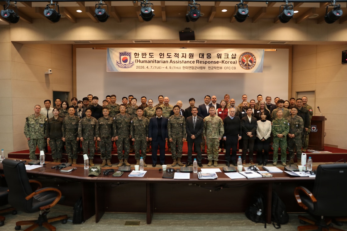 A large group of military personnel and civilians pose for a group photo in a conference hall. They are standing on a stage in front of a large banner that reads "Humanitarian Assistance Response-Korea" with the dates "2026. 4. 7.(TUE) ~ 4. 9. (THU)"
