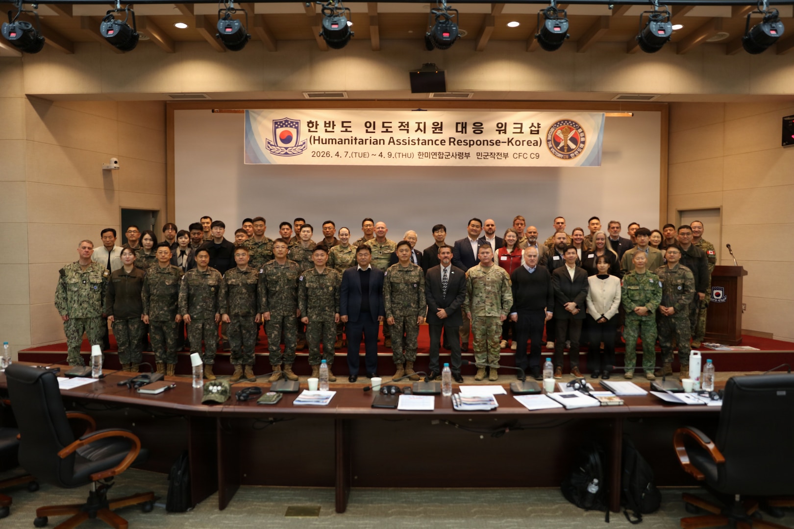 A large group of military personnel and civilians pose for a group photo in a conference hall. They are standing on a stage in front of a large banner that reads "Humanitarian Assistance Response-Korea" with the dates "2026. 4. 7.(TUE) ~ 4. 9. (THU)"