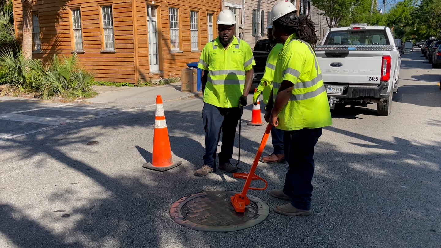 A group of workers wearing safety vests and helmets with one holding an orange tool, standing in a road.