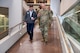 Two men, one in uniform, walking down a large indoor ramp. A group of men in uniform walk behind.