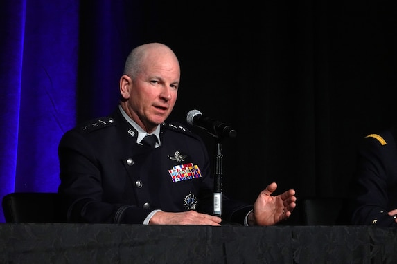 A uniformed man gestures as he speaks seated at a table on a spotlit stage with an affixed microphone