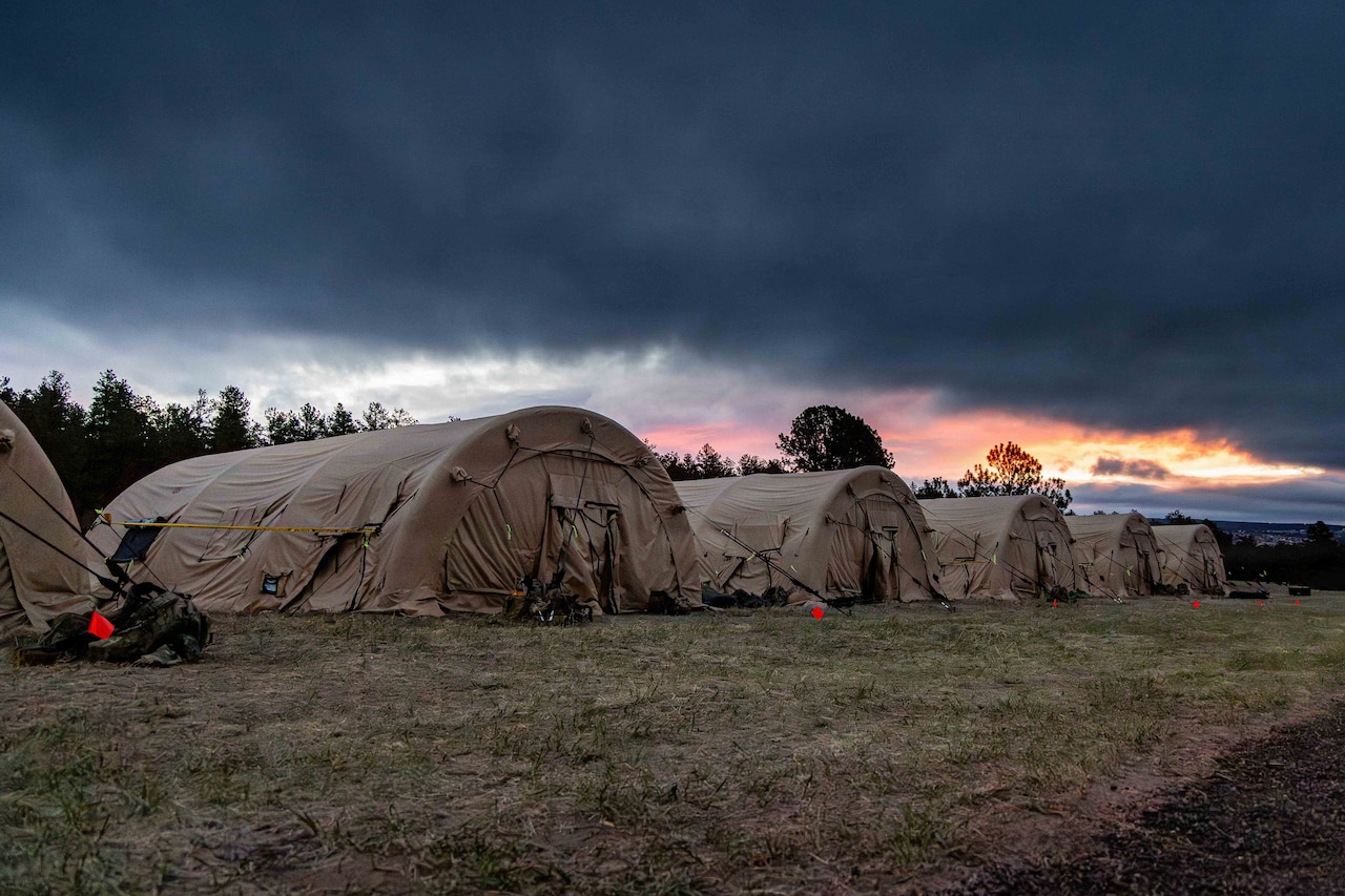 Six military tents are set up in a line on a grassy field under clouds, as the sun sets in the distance.