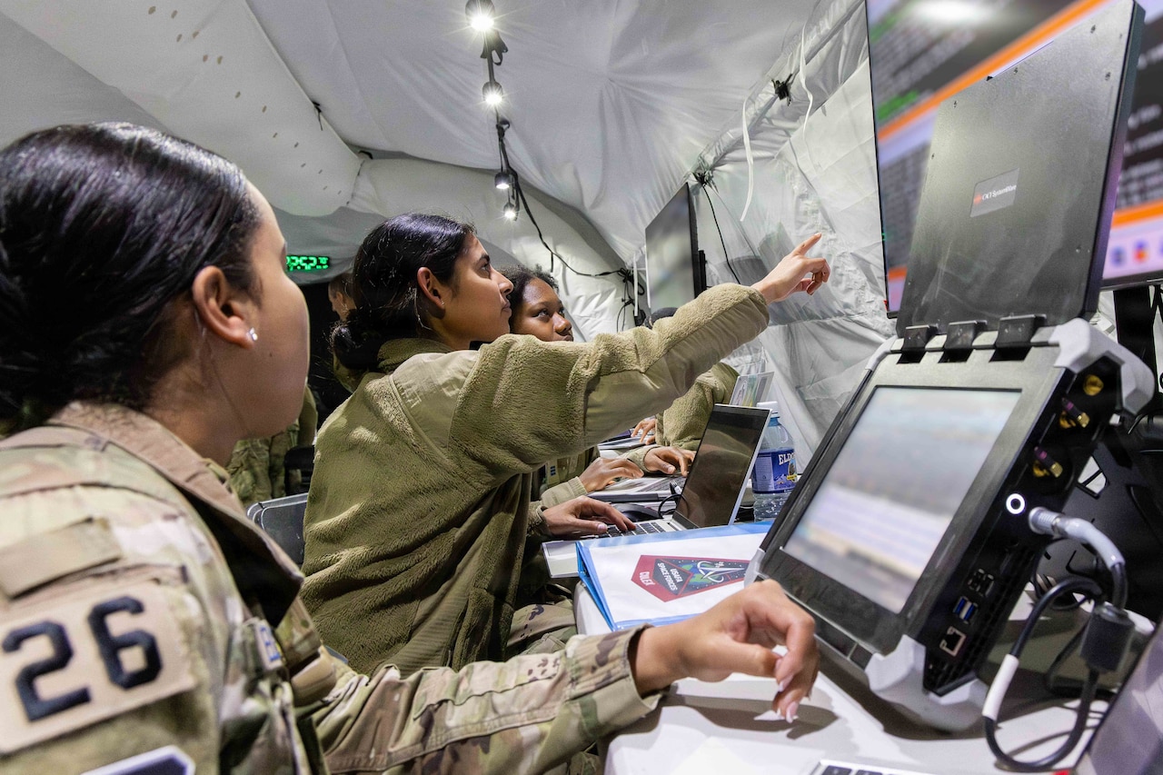 Three women in camouflage military uniforms sit in front of computers and monitors inside a tent; the middle one is pointing to a monitor.