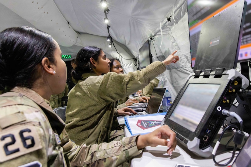 Three women in camouflage military uniforms sit in front of computers and monitors inside a tent; the middle one is pointing to a monitor.