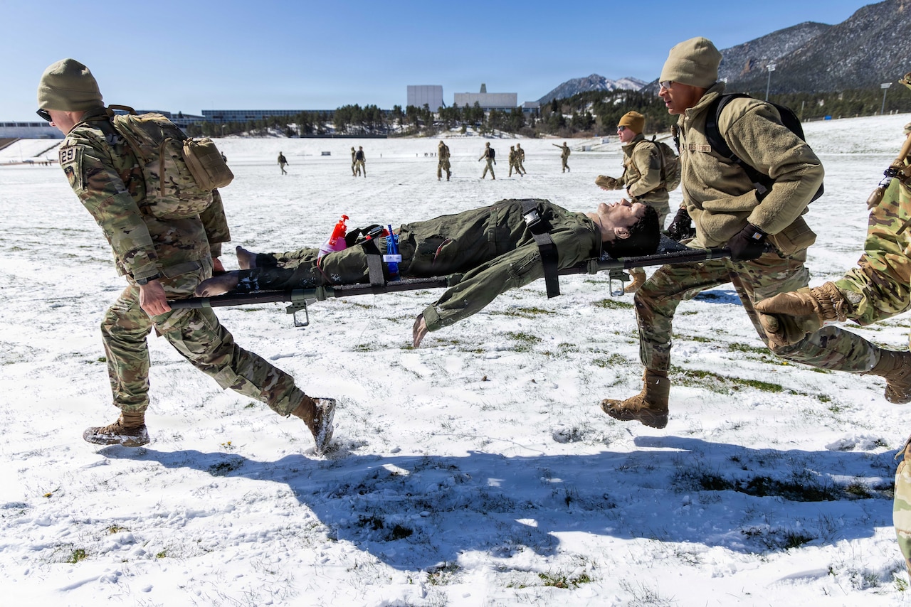 Two people dressed in camouflage military uniforms run outside in the snow while carrying a manikin on a stretcher; nearly a dozen people in similar attire are also running. In the distance are trees, mountains and buildings.