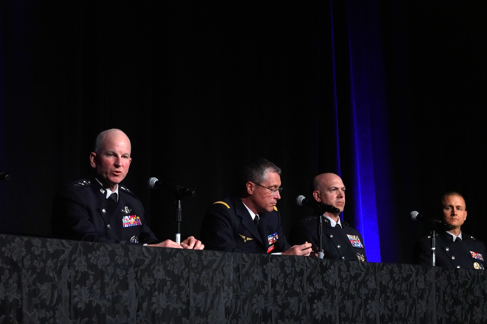 Four uniformed men sit at a table on a spotlit stage with affixed microphones