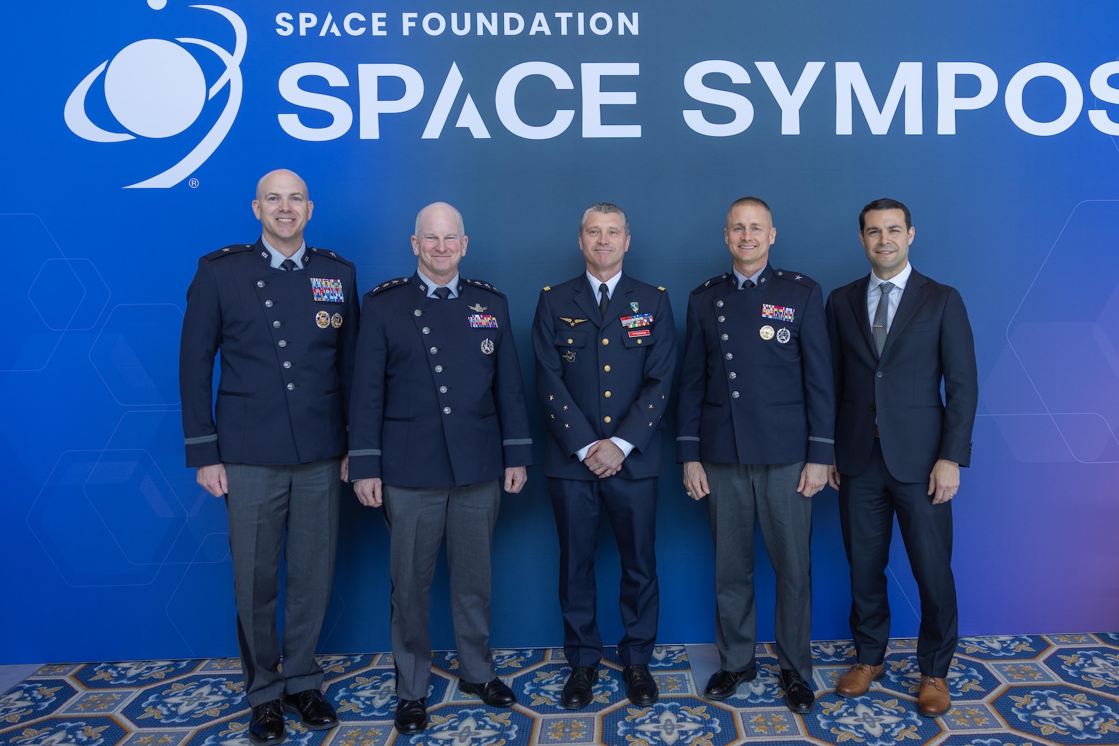 Four uniformed men stand alongside a man in a suit in front of a Space Symposium-branded backdrop