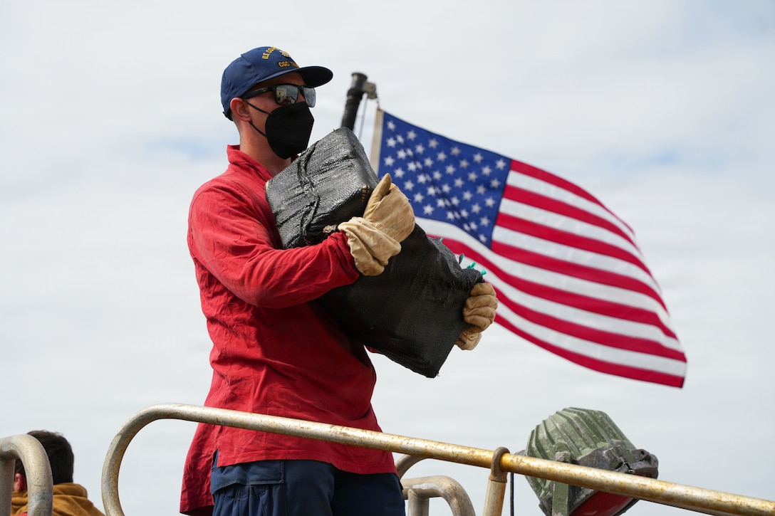 A person wearing a black mask and gloves holds a package wrapped in a black covering outside, with an American flag in the background.