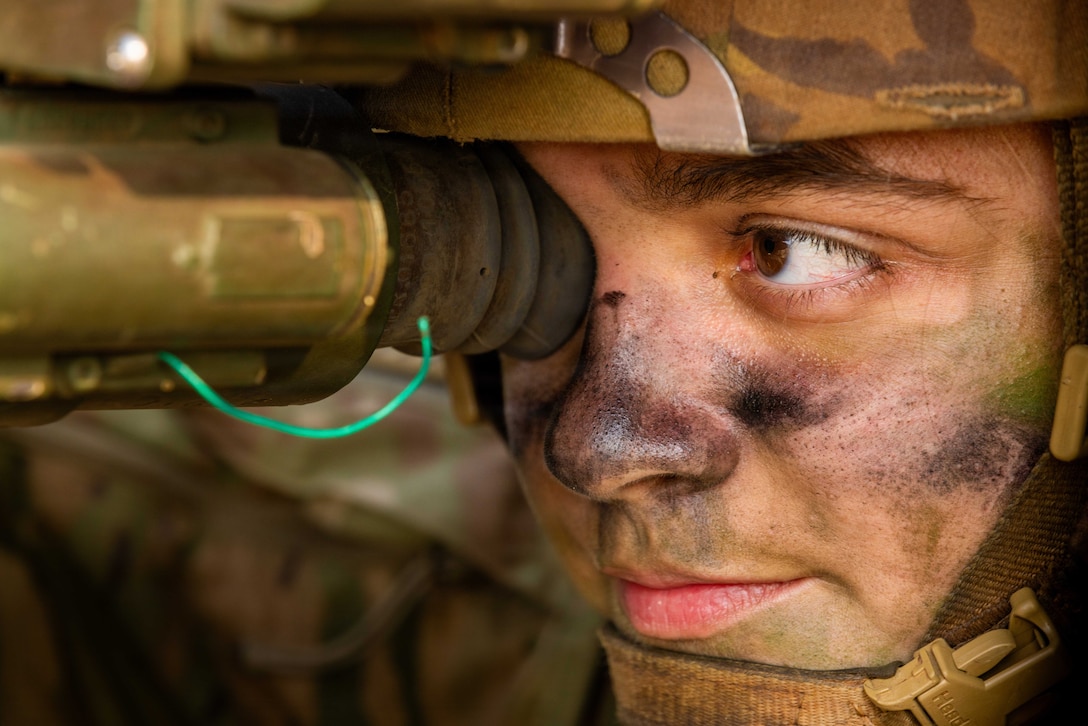 A person wearing a camouflage helmet and black face paint on their nose and cheeks looks through a telescope-like instrument with one eye.