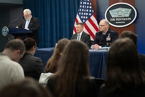 Two men, one in a suit and another in a military dress uniform, sit at a table, while another man in a suit stands at a lectern; several people sitting in chairs looking at the men at the table are shown in the foreground.