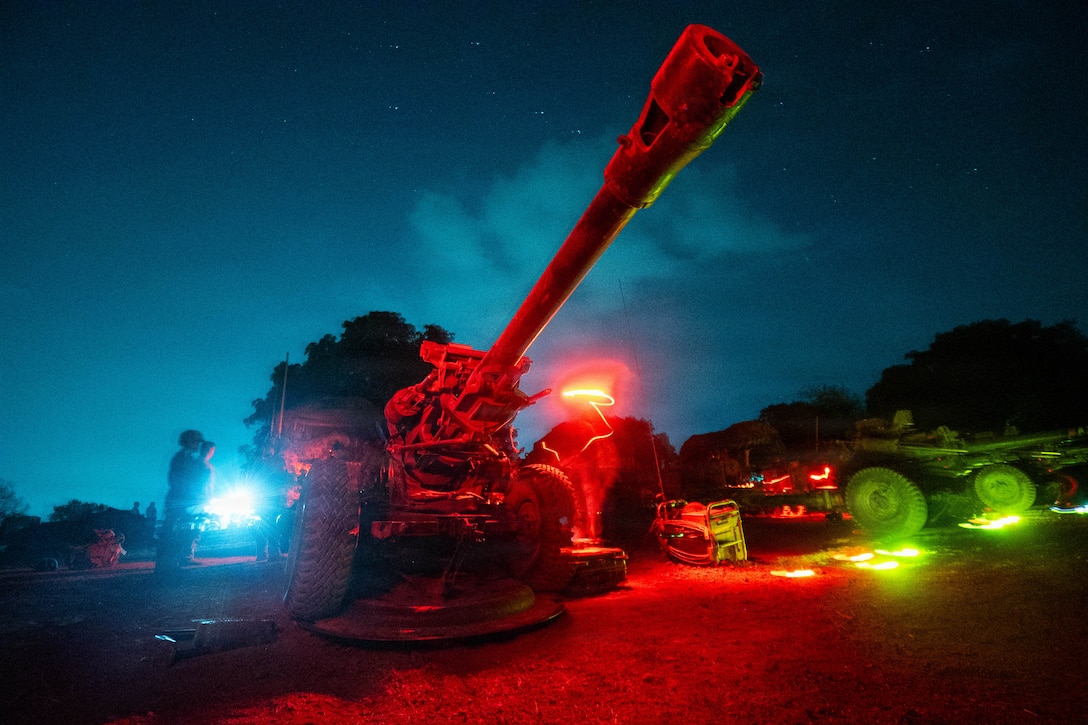 People stand by artillery outside, with red and white lights shining and a dark blue sky and trees in the background.