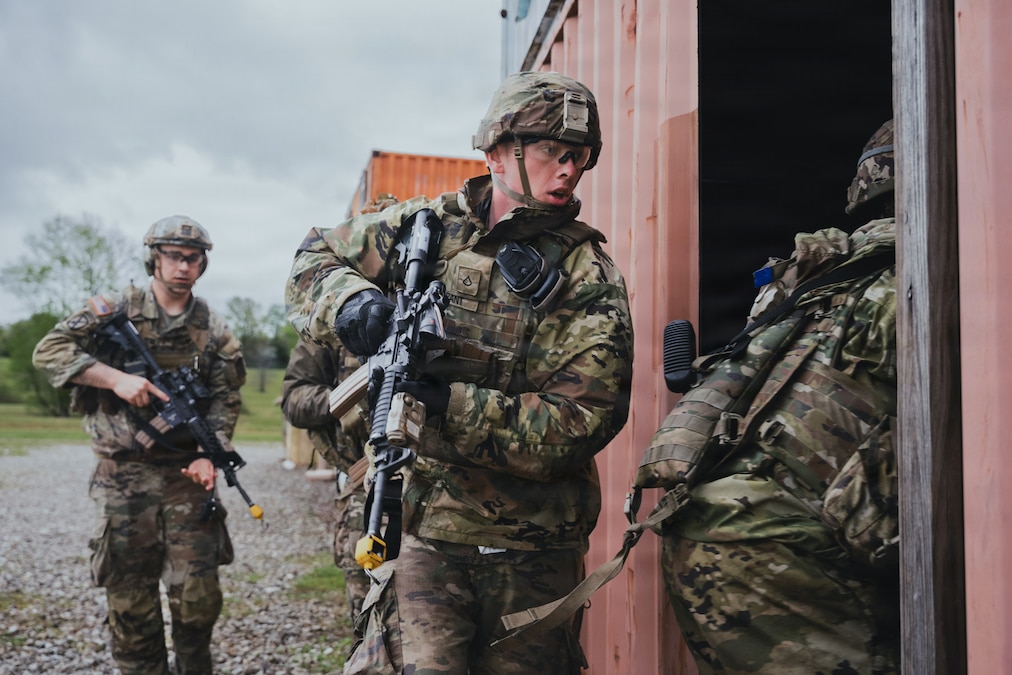 Three soldiers in camouflaged uniforms move into a building holding weapons.