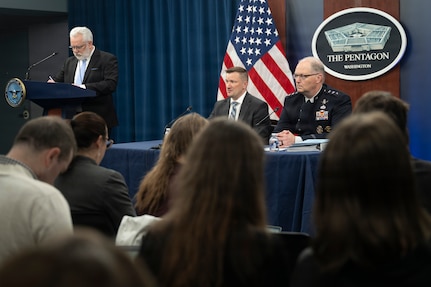 Two men, one in a suit and another in a military dress uniform, sit at a table, while another man in a suit stands at a lectern; several people sitting in chairs looking at the men at the table are shown in the foreground.