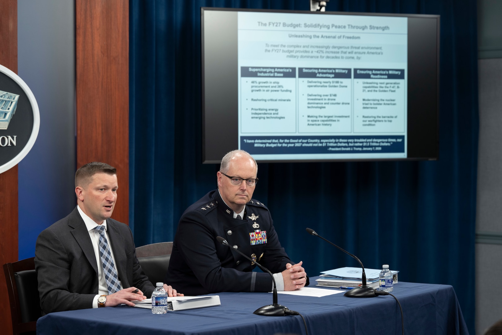 Two men, one in a suit and another in a military dress uniform, sit at a table. Behind them is a display screen showing a slide.