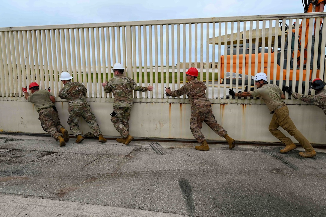 Six airmen wearing hard hats push a metal fence.