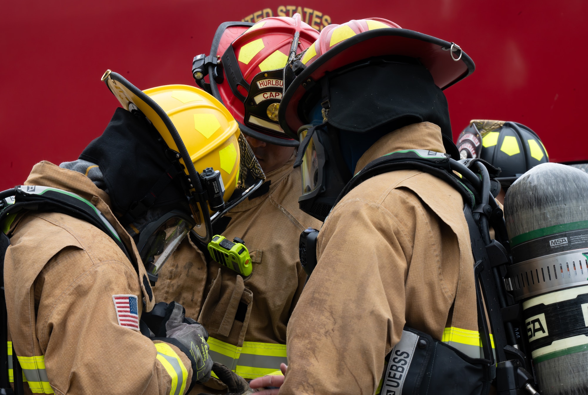 Firefighters in full gear huddle closely to inspect equipment in front of a fire truck.