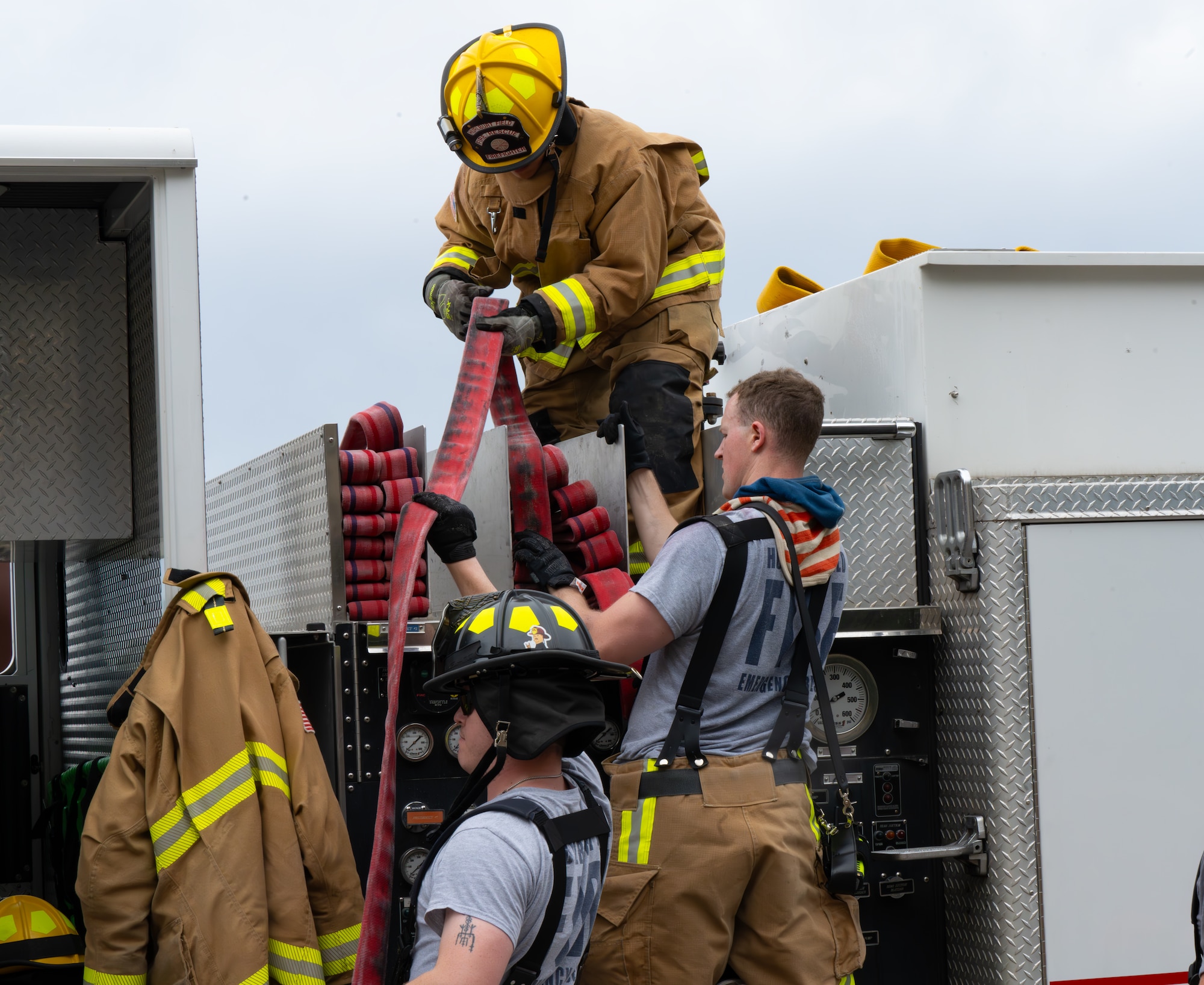Firefighters on a truck rerack hose, with one in full gear guiding the line while two others assist.