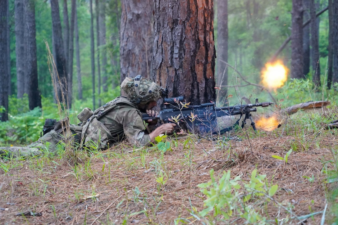 A soldier fires a machine gun while on the ground near a tree, with other trees in the background.