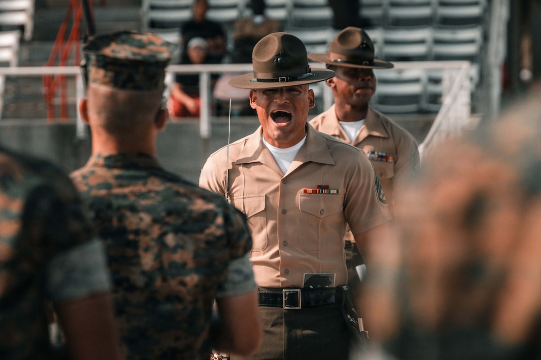 A drill instructor stands in front of people in camouflage uniforms outside, with another instructor and people in stands in the background.
