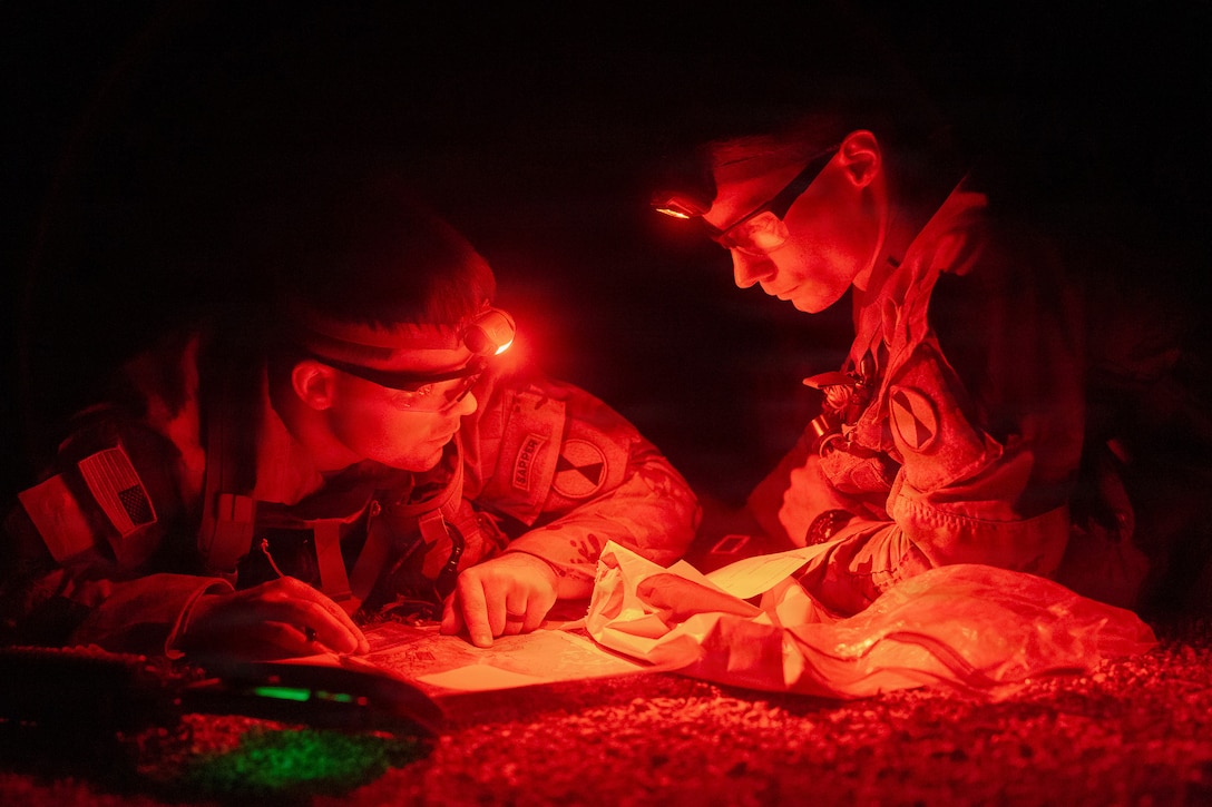 Two soldiers wearing headlamps lean forward to look at papers.