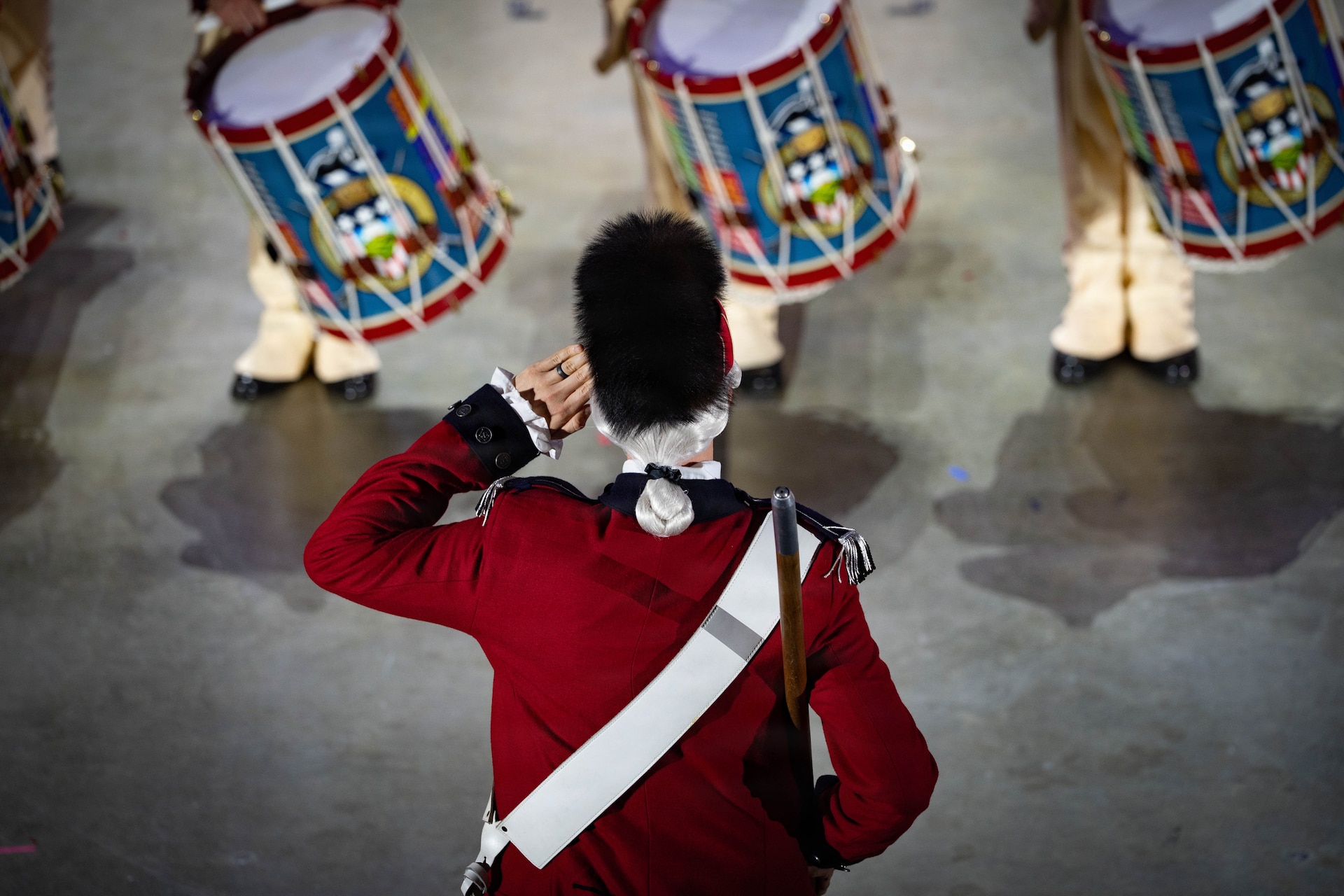An Army soldier wearing a red Revolutionary War-era uniform with a large furry hat is saluting towards a row of drummers. Their drums are adorned with bright colors.