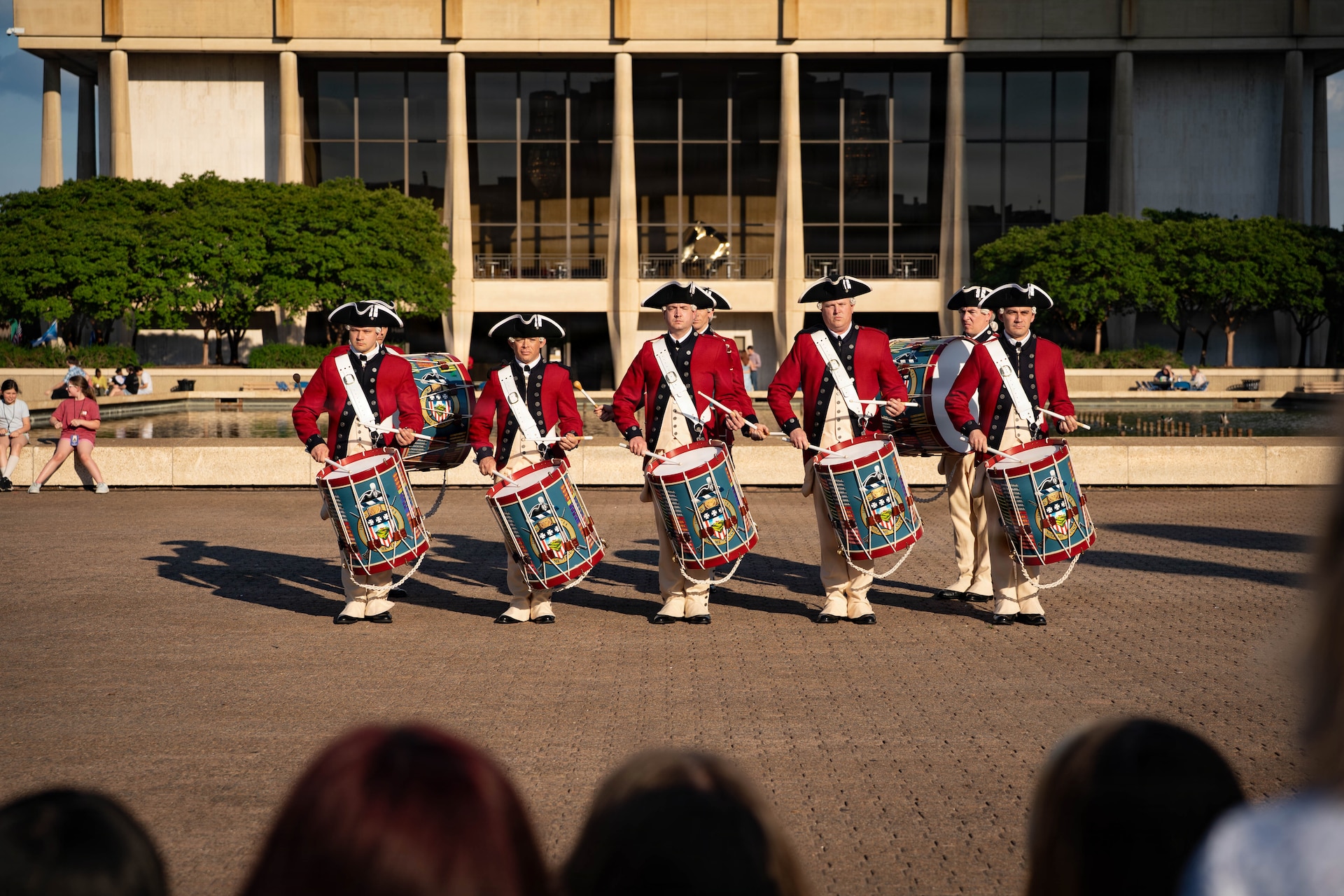 A row of drummers dressed in red Revolutionary War-era uniforms is performing during an outdoor event in front of a large building.