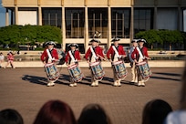 A row of drummers dressed in red Revolutionary War-era uniforms is performing during an outdoor event in front of a large building.