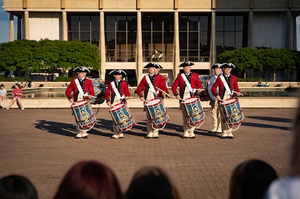 A row of drummers dressed in red Revolutionary War-era uniforms is performing during an outdoor event in front of a large building.