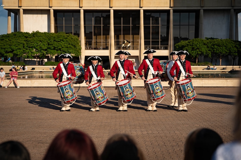 A row of drummers dressed in red Revolutionary War-era uniforms is performing during an outdoor event in front of a large building.