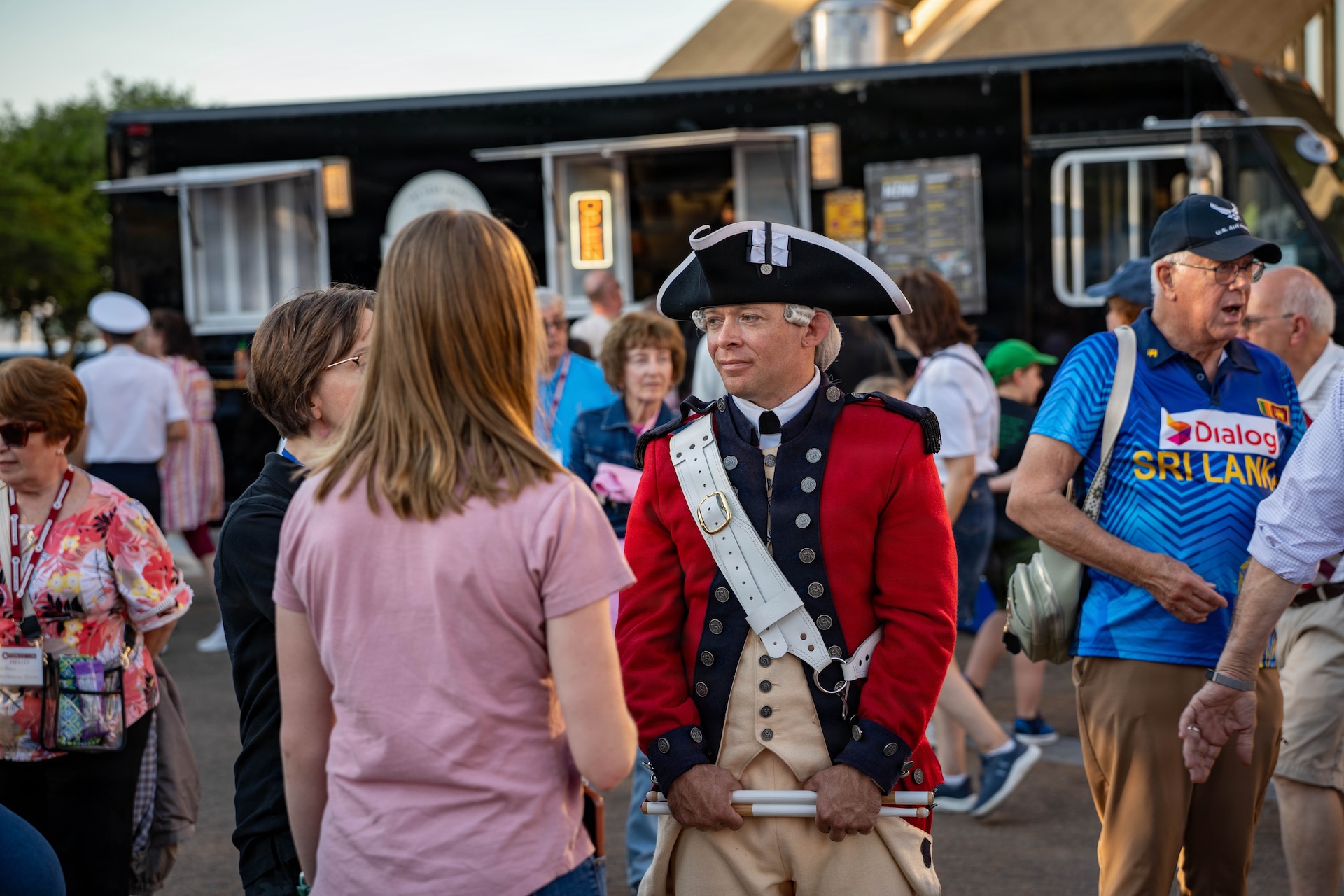 An Army Soldier dressed in a red Revolutionary War-era uniform with dark tri-cornered hat is talking with people during an outdoor event.