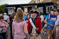 An Army Soldier dressed in a red Revolutionary War-era uniform with dark tri-cornered hat is talking with people during an outdoor event.