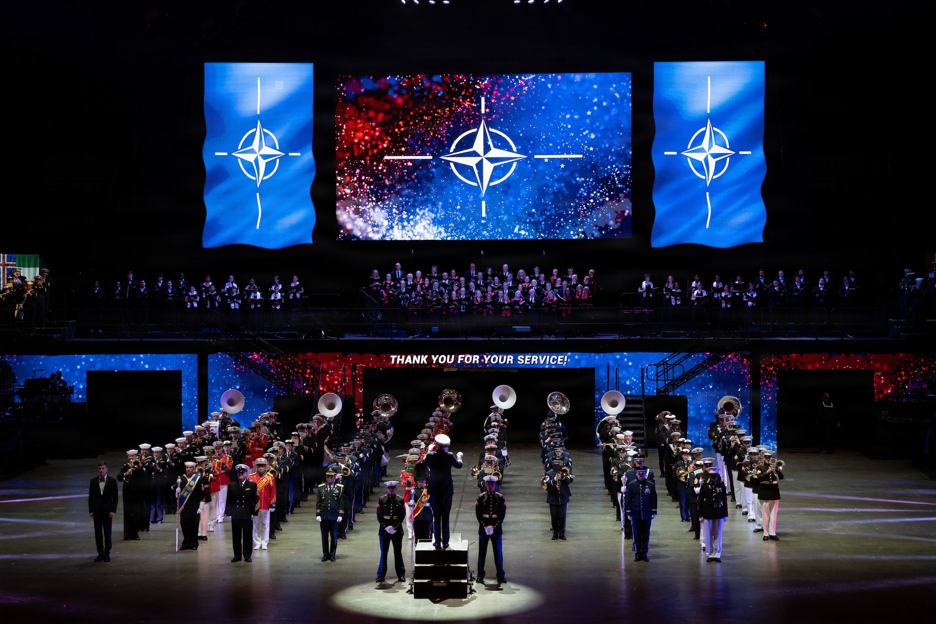 Dozens of instrumentalists wearing a variety of band and military uniforms are standing in rows in formation in a large arena performing in front of large screens that are projecting an image that looks like a compass.