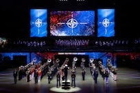 Dozens of instrumentalists wearing a variety of band and military uniforms are standing in rows in formation in a large arena performing in front of large screens that are projecting an image that looks like a compass.