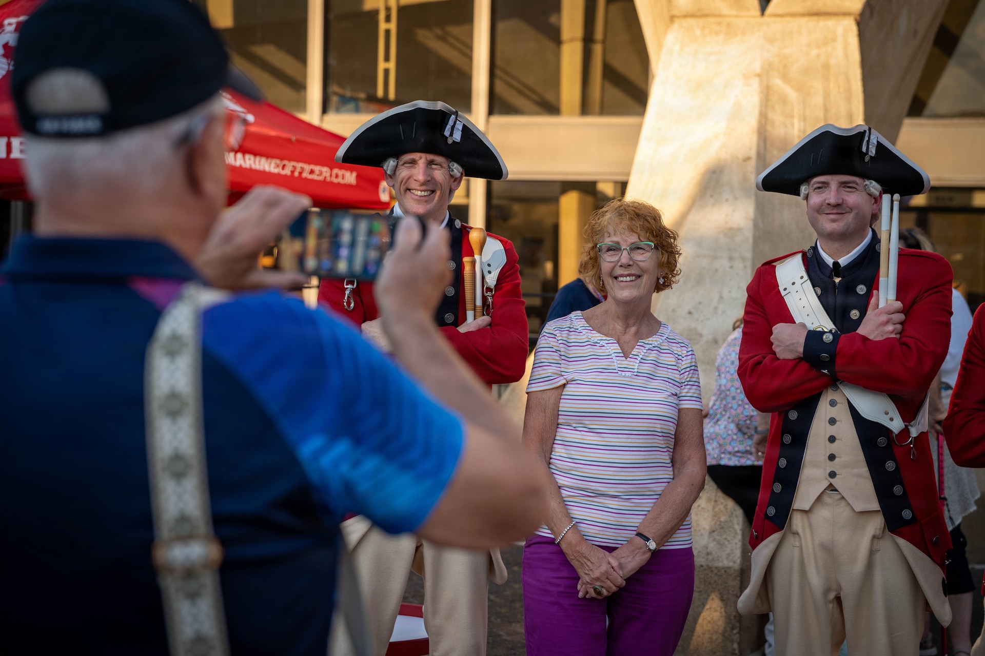 Two Army soldiers in red Revolutionary War-era uniforms are standing on either side of a woman who is posing for a picture being taking by a man in the foreground.