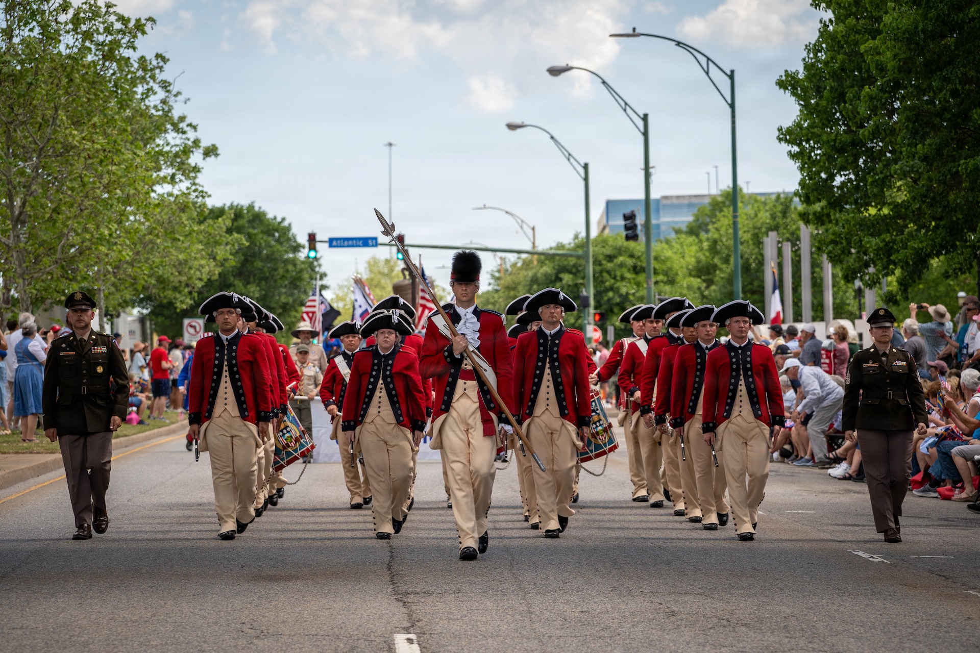 A military band dressed in red Revolutionary War-era uniforms in marching in formation in the middle of a street during a parade, heading toward the camera. There are crowds on the sidewalks, and they are flanked by one soldier in green service dress on either side of the band.