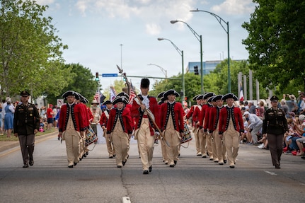 A military band dressed in red Revolutionary War-era uniforms in marching in formation in the middle of a street during a parade, heading toward the camera. There are crowds on the sidewalks, and they are flanked by one soldier in green service dress on either side of the band.