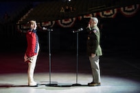 Two members of the military are raising their right hands while facing each other on a stage lit by spotlights. The soldier on the left is wearing a red Revolutionary War-era uniform, and the soldier on the right is wearing a  modern green service uniform.