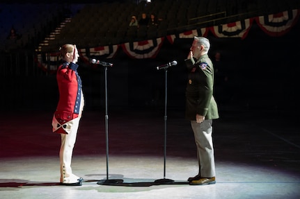 Two members of the military are raising their right hands while facing each other on a stage lit by spotlights. The soldier on the left is wearing a red Revolutionary War-era uniform, and the soldier on the right is wearing a  modern green service uniform.