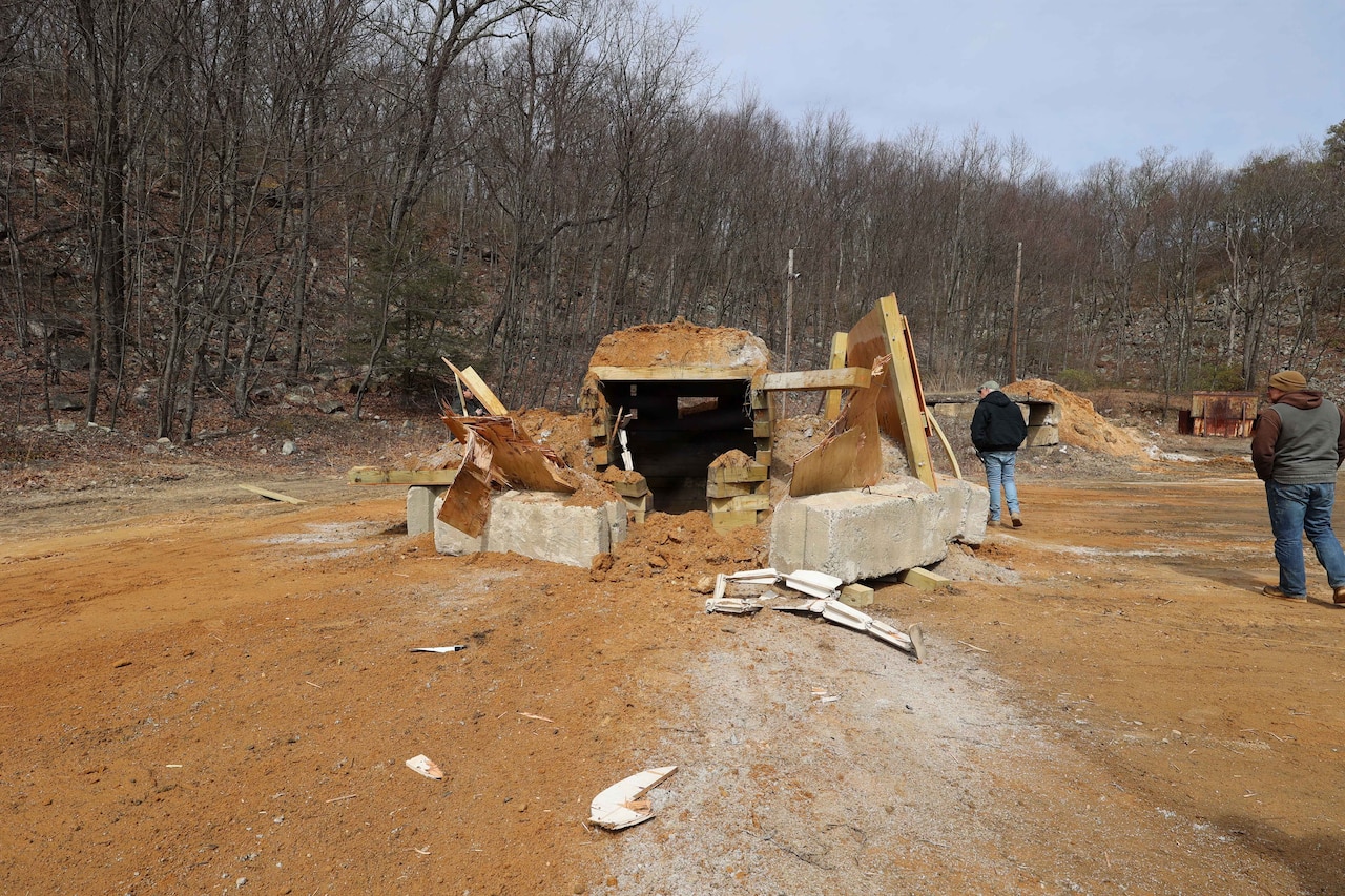 Two people in casual attire observe the remains of a partially destroyed bunker in a dirt terrain with trees in the background.