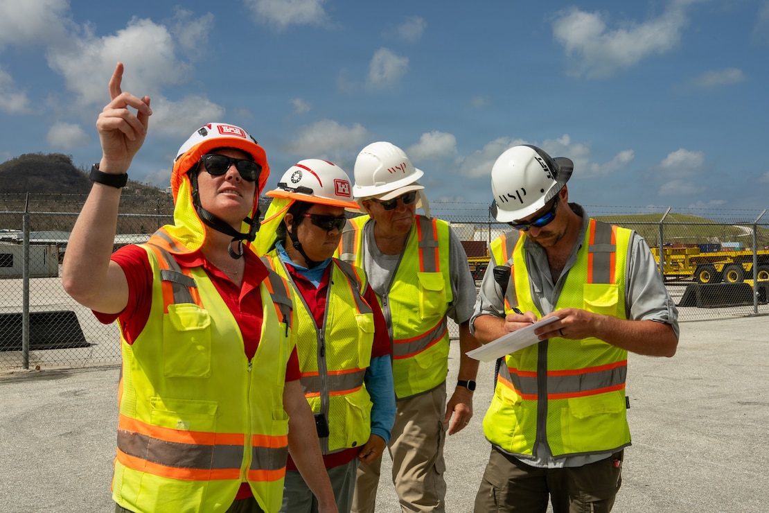 Four individuals in hard hats and safety vests stand next to each other. One person is writing on a clipboard.