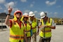 Four individuals in hard hats and safety vests stand next to each other. One person is writing on a clipboard.
