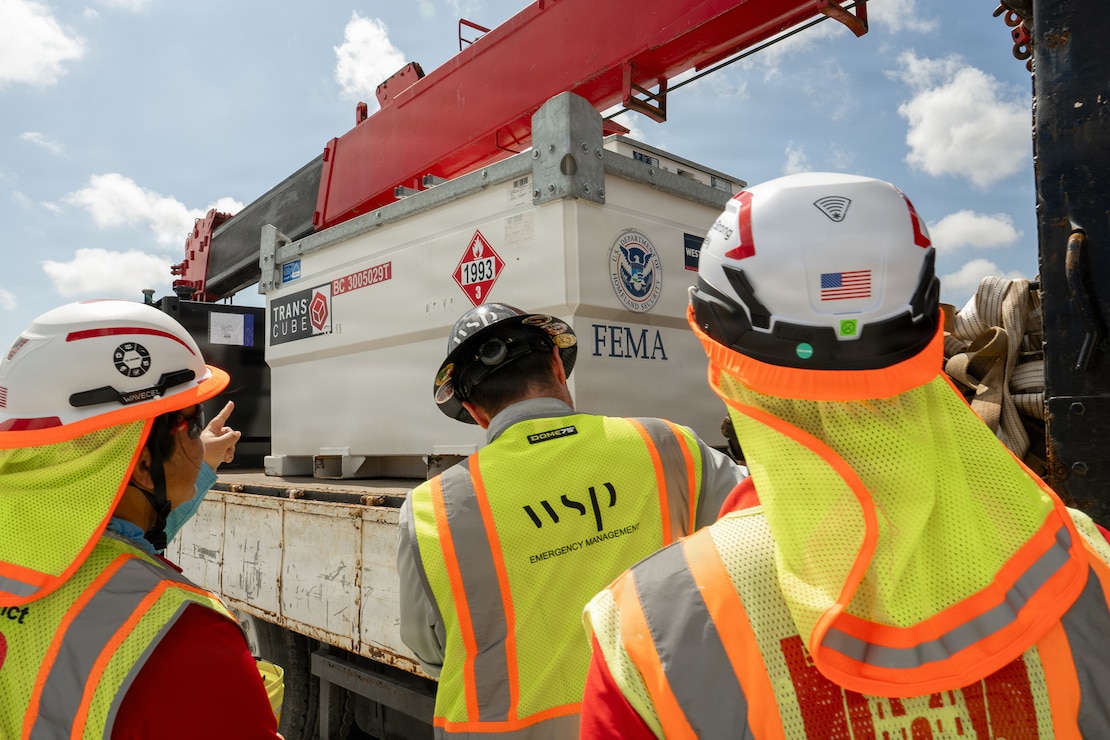 Three individuals look at a generator which is sitting on a flatbed truck.