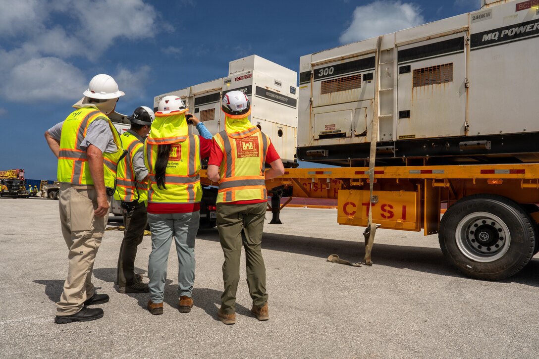 A group of individuals in hard hats and safety vests stand next to a flatbed truck loaded with large generators.